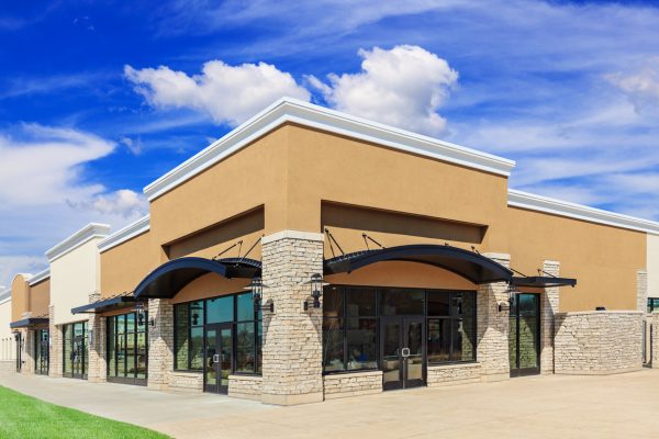 New commercial development featuring a street view of a Strip Mall with green grass, sidewalk and patio space. Blue sky and clouds are in the background.