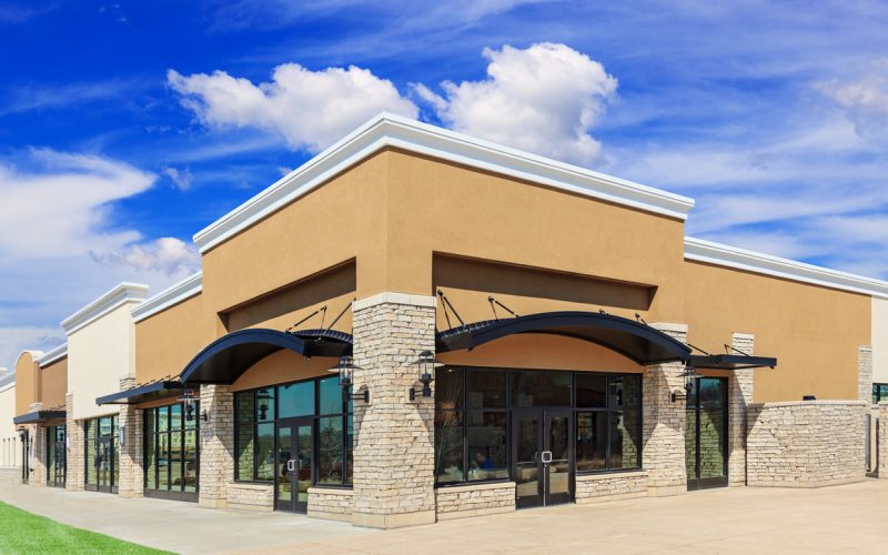 New commercial development featuring a street view of a Strip Mall with green grass, sidewalk and patio space. Blue sky and clouds are in the background.